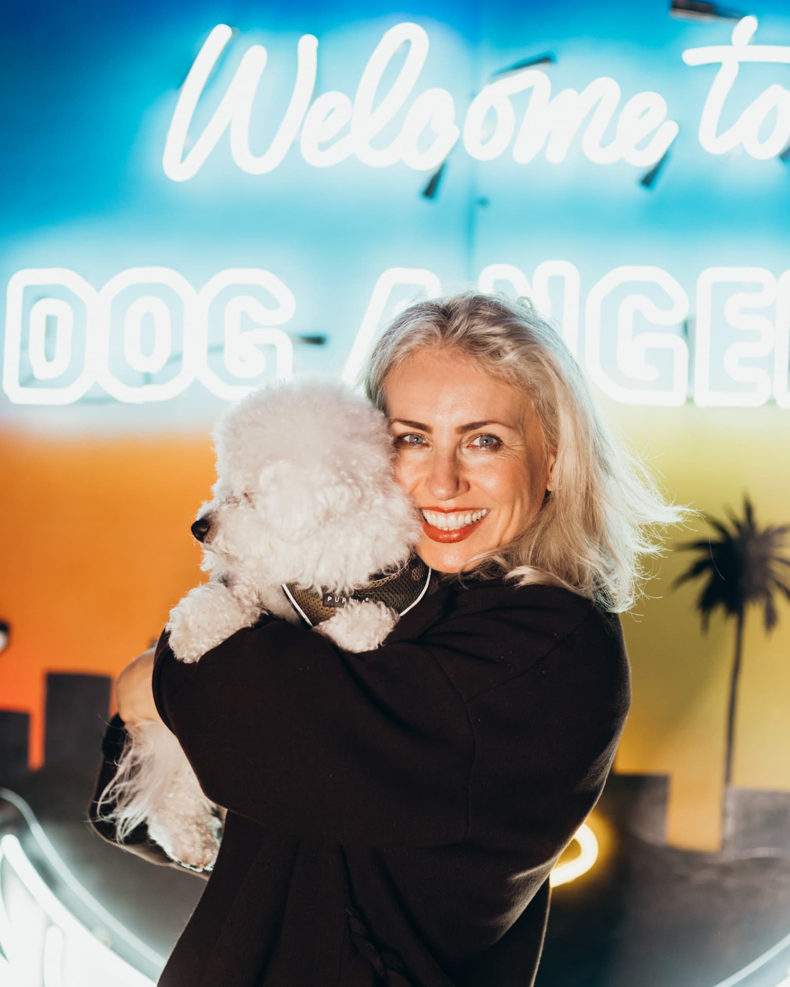Pet parent hugging her bichon in front of the 'Welcome to Dog Angeles' neon